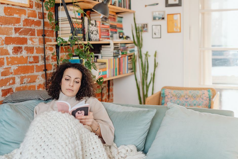 Mujer leyendo un libro tumbada en el sofá bajo una manta gruesa de punto