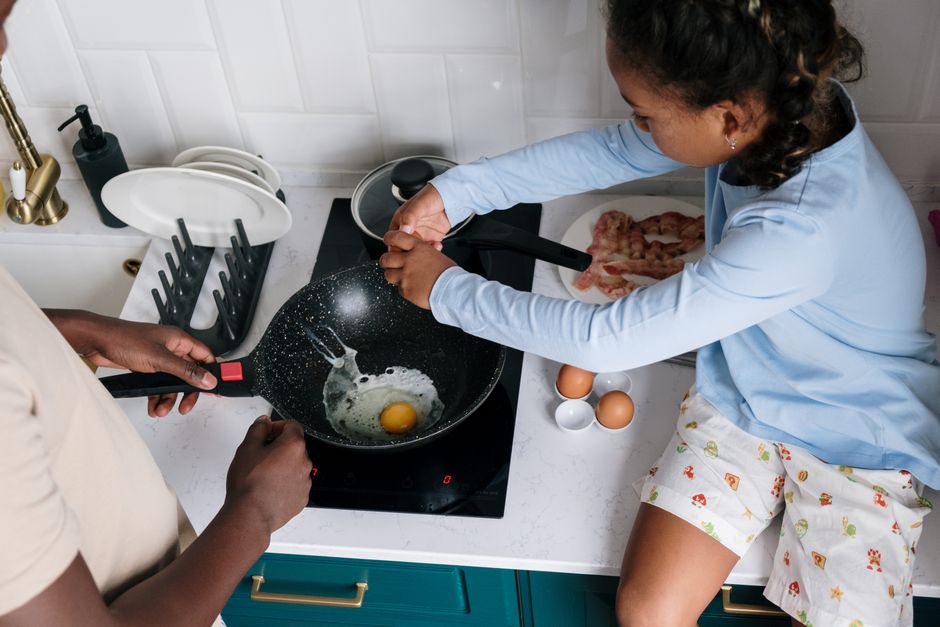 Una niña y un adulto cocinando un huevo frito en una sartén sobre una placa eléctrica negra